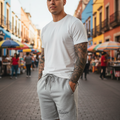 A man standing on a cobblestone street wearing a white crew-neck t-shirt and light gray drawstring sweat shorts with a small script logo on the left leg. The background shows a colorful, blurred outdoor market scene with buildings and stalls.
