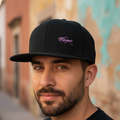 A man wearing a black baseball cap with a purple embroidered script logo on the front, set against a blurred background of colorful building walls.