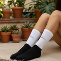 A person seated on a patio wearing black crew socks with white ribbed cuffs and small script branding on the cuffs. The background features potted green plants and a beige stone floor.
