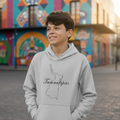 A smiling young man wearing a light gray hoodie with a black outline map of Newfoundland and the word 'Tamaulipas' printed on the chest, standing outdoors in front of a brightly painted mural building.