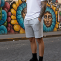A man wearing a white crew-neck t-shirt and light gray shorts stands on a cobblestone street in front of a colorful graffiti wall.