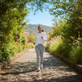 A woman jogging on a cobblestone path surrounded by lush green trees and bushes under a clear blue sky.