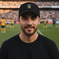 A man wearing a black crewneck t-shirt and a black Tigres UANL Twill baseball cap with a small crest logo, standing in a stadium with a soccer field and crowd in the background.