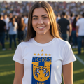 A smiling young woman stands on a football field wearing a white T-shirt with a large blue and yellow Tigres UANL graphic on the front, with a blurred crowd and stadium in the background.