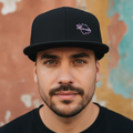 A man wearing a black snapback cap with a small purple embroidered logo that reads 'Hermana' against a textured, multicolored wall background.