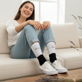 A woman sitting on a cream-colored couch wearing white crew socks with black lower sections and white low-top sneakers. The socks have Tlaxcala cursive brand name printed on them.
