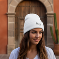 A woman with long brown hair wearing a white Veracruz embroidered beanie with a small black embroidered logo, standing in front of a warm-toned stone archway and wooden door.