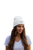A woman with long brown hair wearing a white knit beanie with a small black embroidered logo on the front, paired with a white t-shirt, set against a solid white background.