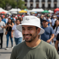 A smiling man in a white bucket hat with a black script logo and an olive green t-shirt, photographed outdoors in a crowded public space with a blurred background.
