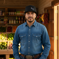 A man wearing a blue denim long-sleeve button-up shirt, dark blue jeans, a brown leather belt with a large silver buckle, and a black baseball cap stands in front of a rustic wooden wall and shelves of produce and goods.
