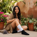 A woman sits on a patio in a garden setting, wearing a brown linen shirt, brown shorts, white socks with black text, and black socks.