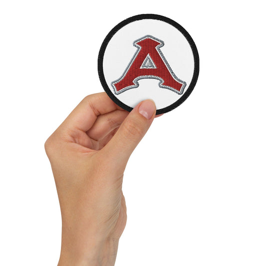 A hand holding a round embroidered patch with a white background and a red letter 'A' in the center, resembling an Acereros de Monclova logo.