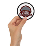 A hand holding a round white embroidered patch with the text 'Algodoneros de UNION LAGUNA' and a number '1914' in the center.