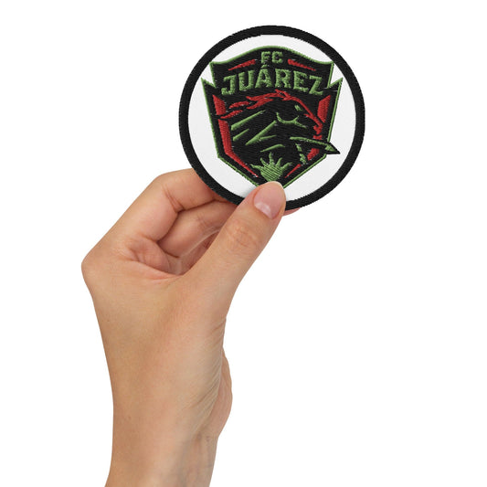 A hand holding a round white embroidered patch with 'FC JUAREZ' logo in red and green.
