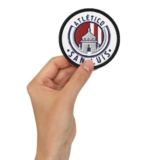 A hand holding a white round embroidered patch with a Club Atletico logo.