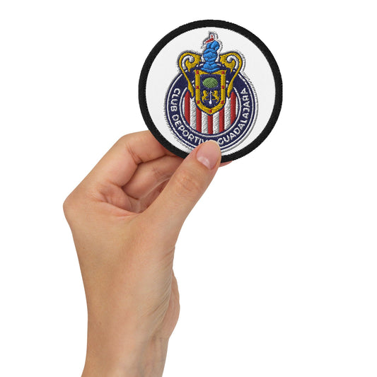 A hand holding a round white embroidered Guadalajara Chivas patch with a colorful emblem.