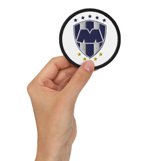 A hand holding a white embroidered patch with a white Club Monterrey logo and stars.