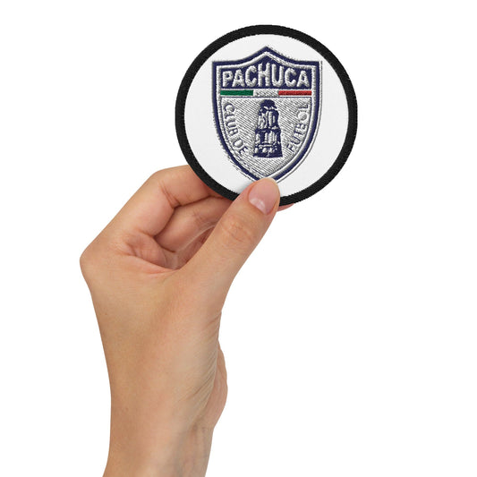 A hand holding a round white embroidered patch with the text 'PACHUCA' and a crest.