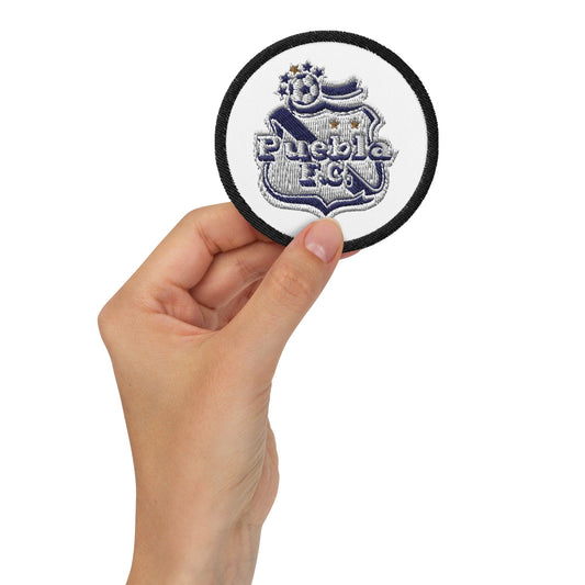 A hand holding a wjote embroidered patch with the text 'PUEBLA FC' on it.