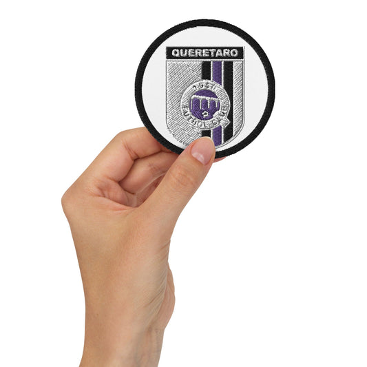A hand holding a round white embroidered patch with the text 'QUERETARO' and a emblem in the center.