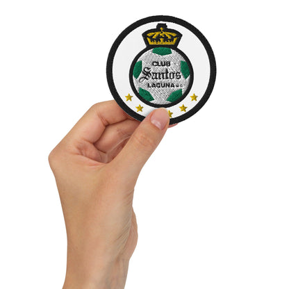 A hand holding a round black embroidered patch with the text 'Club Santos Laguna' and a soccer ball design.