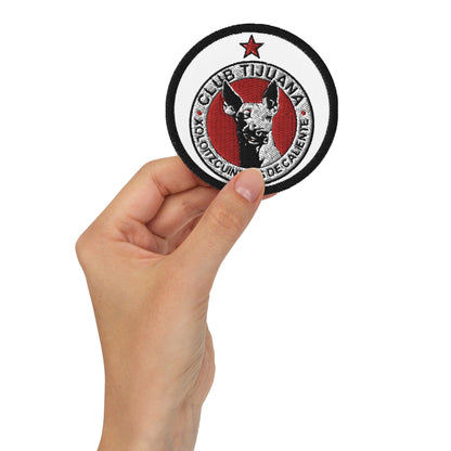 A hand holding a round embroidered patch with a red and white logo and the text 'CLUB TIJUANA'.
