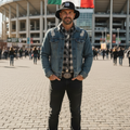 A man wearing a Mariachis de Guadalajara black bucket hat, a distressed blue denim jacket over a plaid shirt, black jeans, and brown boots stands in front of a stadium with a Mexican flag hanging above him.