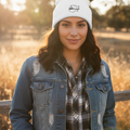 A woman wearing a white beanie with a black embroidered logo, a distressed blue denim jacket, and a black-and-white plaid shirt, standing outdoors in a sunlit field.