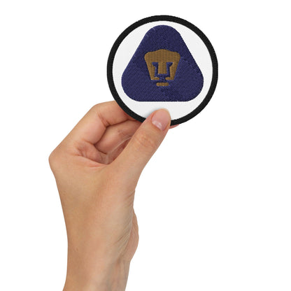A hand holding a round embroidered patch with a white border and a golden puma logo in the center, predominantly blue in color.