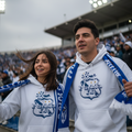 A man and a woman wearing matching white and blue Club Puebla  hoodie sweatshirts and scarves, standing in a stadium crowd.