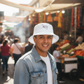 A smiling man wearing a white Sonora bucket hat with a black logo and a light blue denim jacket over a white t-shirt, standing in a sunny outdoor market street.