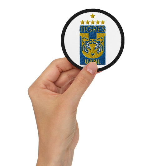 A hand holding a round white embroidered patch featuring the Tigres UANL logo in blue and yellow.