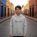 A young boy stands on a cobblestone street wearing a light gray hoodie with 'Yucatán' printed in black script inside a map outline. Colorful buildings line both sides of the street in the background.