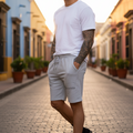 A man wearing a white crewneck t-shirt and light gray drawstring sweat shorts stands on a cobblestone street lined with colorful buildings.