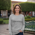 A woman stands outdoors in a park-like setting wearing a light gray long-sleeve shirt with a black outline graphic of the state of Yucatán and the word 'Yucatán' across the front, paired with dark blue jeans.