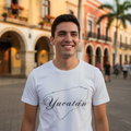 A smiling man wearing a white t-shirt with a black outline of the state of Yucatan and the word 'Yucatán' written across the front, standing in a sunny outdoor street with colonial-style buildings and palm trees in the background.