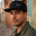 A man wearing a black baseball cap with a purple embroidered logo that reads 'Juarache' and an olive green shirt, standing outdoors against a blurred background of colorful buildings.