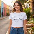 A woman stands on a colorful outdoor street wearing a white cropped T-shirt with a black outline of Yucatán and the word 'Yucatán' printed across the chest, paired with blue jeans.