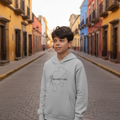A young boy stands on a cobblestone street wearing a light gray hoodie with a black graphic of the state of Mexico and the word 'Zacatecas' written across the chest, with colorful colonial-style buildings lining the street in the background.