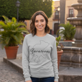 A woman stands outdoors in a plaza wearing a light gray long-sleeve crewneck sweatshirt with a black outline map of Zacatecas and the word 'Zacatecas' printed across the chest, paired with dark blue jeans.