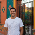 A smiling man wearing a white graphic t-shirt with a black outline map of Yucatecos and the word 'Yucatecas' in script, standing in front of a colorful mural wall and an arched black metal gate.