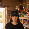 A woman wearing a black baseball cap with a small white embroidered logo, standing in a rustic shop interior with shelves of snacks and limes in the background.