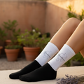 A person's legs wearing black and white crew socks with a butterfly graphic, sitting on a beige stone ledge with potted plants in the background.