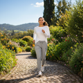 A woman jogging on a cobblestone path in a lush green garden, wearing a white long-sleeve top, light gray drawstring sweatpants, and white sneakers.