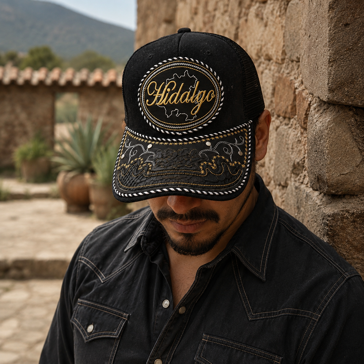 A Mexican model wearing a black Hidalgo charro-style hat in the Mexican countryside near stone walls. The embroidered Hidalgo name and map design are clearly visible on the front.