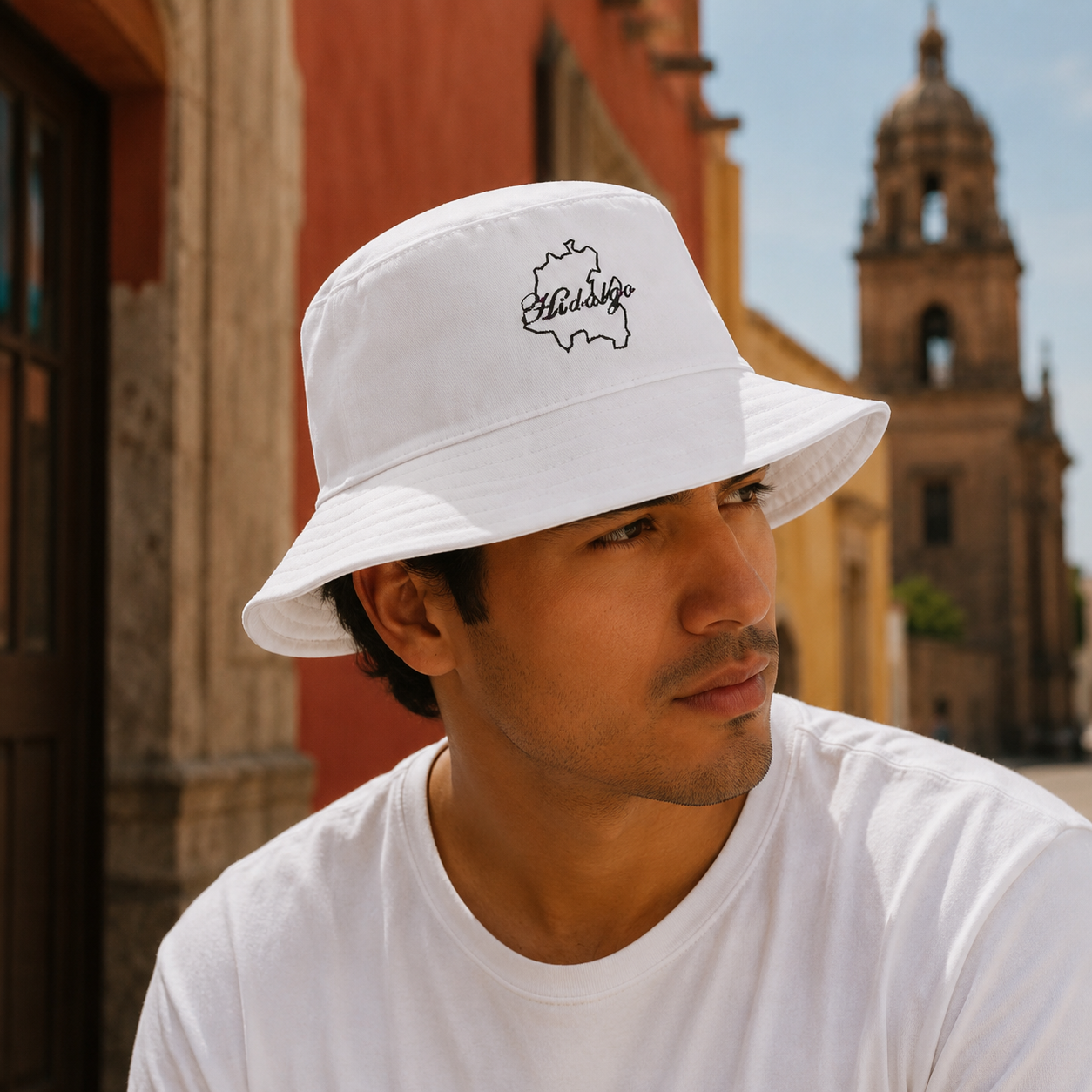 A man wearing a white bucket hat with a black outline graphic of the state of Hidalgo and the word 'Hidalgo' written in script, photographed outdoors in front of a warm-toned building and a church tower.