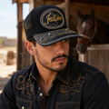 A Mexican model wearing a black Jalisco charro-style hat at a charro ranch near a horse stable. The embroidered Jalisco name and map design are clearly visible on the front.