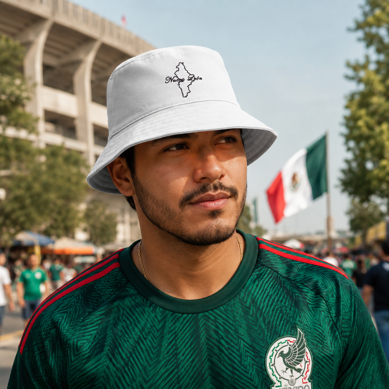 A man wearing a white Nuevo Leon bucket hat with a black outline logo and a green Mexico soccer jersey, photographed outdoors in front of a stadium and flags.