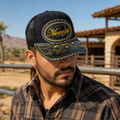 Mexican man wearing a black embroidered “Mexicali” trucker hat at a desert ranch with mountains in the background.