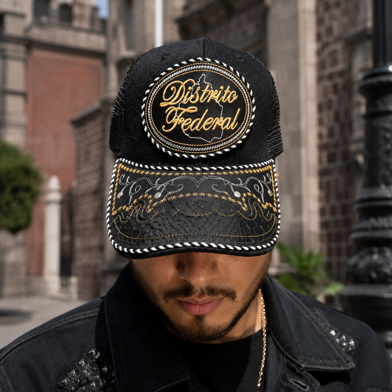 A Mexican model wearing a black Mexico City charro-style hat in a historic urban neighborhood. The embroidered Mexico City name and map design are clearly visible on the front.